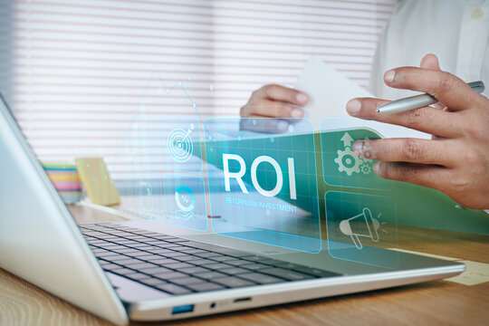 Business professional reviewing document on desk with laptop and holographic ROI return on investment icon, concept of business growth, profitability analysis, and financial return.