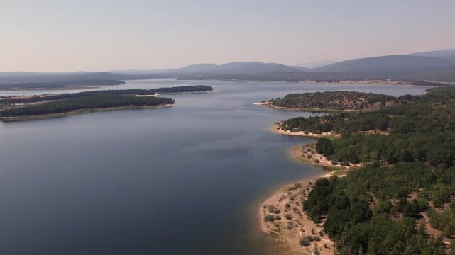 Drone flying over La Cuerda del Pozo Reservoir in Soria with a hazy mountain horizon and pine forests on the shoreline