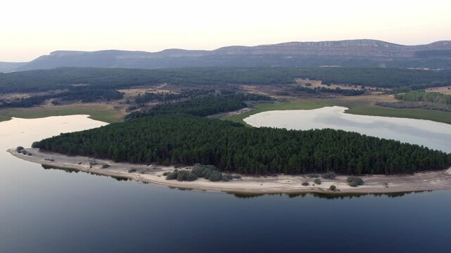 Drone flying over La Cuerda del Pozo Reservoir revealing a beautiful pine forest and mountains at sunset in Soria, Spain