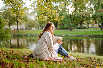 Fototapeta premium Young woman sits on grass in park with cup of hot coffee at sunset. Woman in coat enjoys autumn day outdoors. Concept of style, fashion, and leisure.