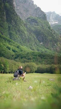 Female hiker using a smartphone while resting in a meadow during a hiking trip with a scenic mountain view, combining outdoor adventure, navigation and digital connectivity.