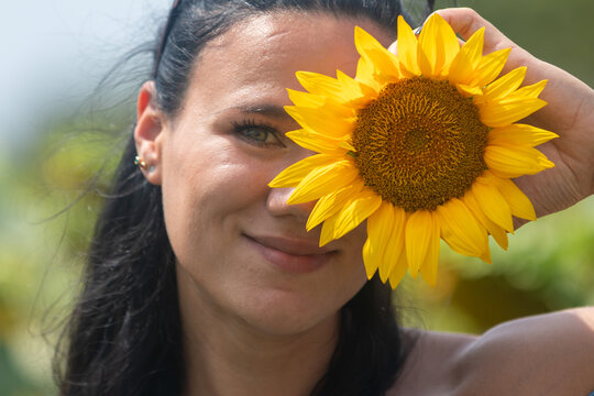 Smiling woman holding a bright yellow sunflower to her face outdoors, creating a cheerful and vibrant summer vibe.