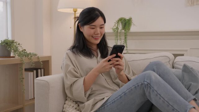 Asian woman using smartphone sofa bright living room home, smiling during casual mobile communication, relaxing indoors with houseplant, soft daylight, neutral decor, comfortable furnishings and calm