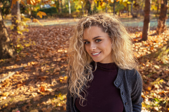 Charismatic cute young blonde woman with curly natural beautiful hair standing while looking at camera , autumn golden hour outdoors portrait high angle view showing dry leaves on the ground