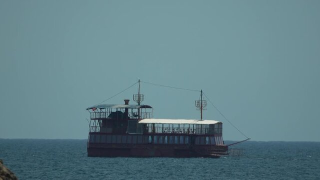 Tourist ferry boat cruises across blue ocean waters near a rocky coastline under clear skies. Panning shot following the moving vessel.