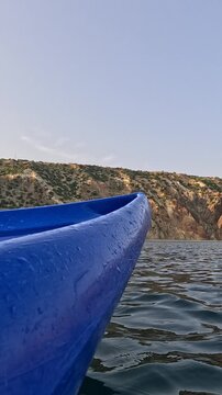 Blue kayak bow glides across calm sea water near high rugged mountains under clear sky. Static low-angle shot.