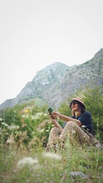 Female hiker using a smartphone while resting in a meadow during a hiking trip with a scenic mountain view, combining outdoor adventure, navigation and digital connectivity.