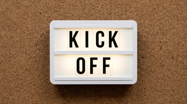 Glowing lightbox sign displays the words Kick Off in black capital letters on a white background mounted on a corkboard for announcements.