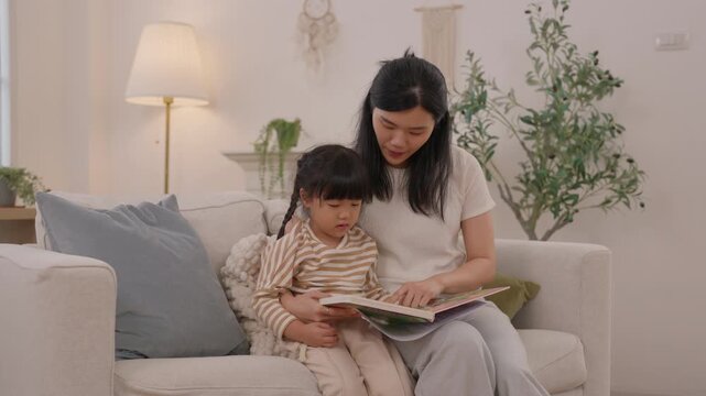 Mother and daughter reading colorful vdo book together on cozy living room sofa, sharing tender family moment with child and woman at home in warm peaceful interior