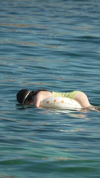 Summer water recreation featuring a woman resting face down on a small inflatable ring in calm ocean waves. Medium static shot.