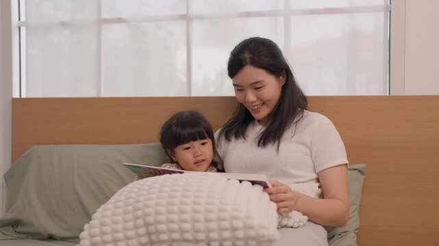 Asian mother and daughter reading storybook together on bed in bright bedroom, warm family moment with child and woman sharing gentle smiles beneath textured blanket at bedtime