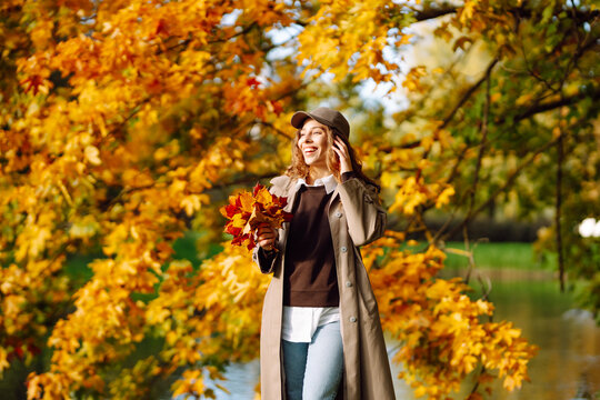 Stylish woman in cap and holding bouquet of fallen leaves relaxes by pond in park. Woman enjoys autumn park, playing with yellow leaves at sunset. Concept is seasonality, style, and relaxation.