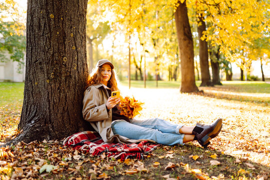 Woman sits on blanket near tree with phone and bouquet of bright yellow leaves. Woman relaxes in autumn park at sunset with smartphone. Concept of seasonality, relaxation. Lifestyle.