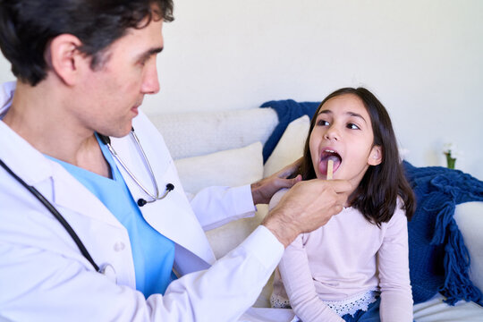 Pediatric house call, doctor examining sick child's throat with a tongue depressor in a bright home living room