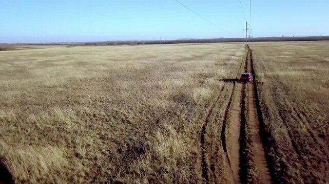 Aerial view of red SUV car driving on dirt road through wide dry steppe field under clear blue sky