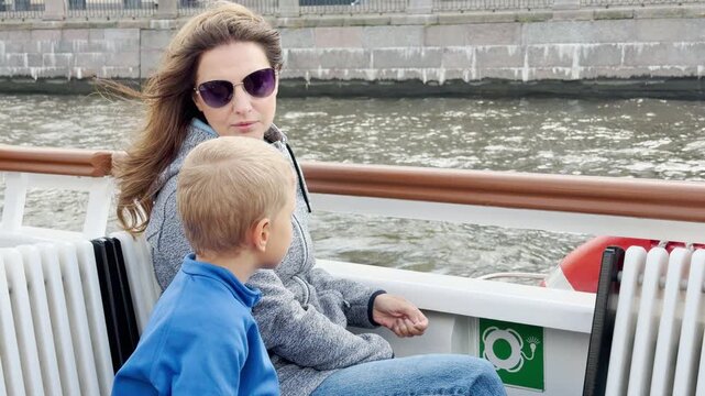 A woman and young boy sit thoughtfully together on a boat deck in Saint Petersburg. They watch the gentle water near a stone wall. This peaceful ride evokes warm calm.