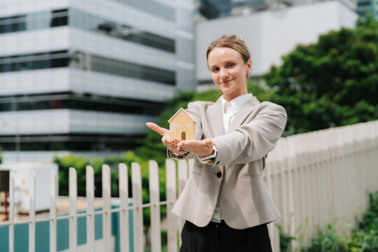 Businesswoman holding small wooden house model on her palm in front of a modern glass office building, real estate investment, property and home ownership