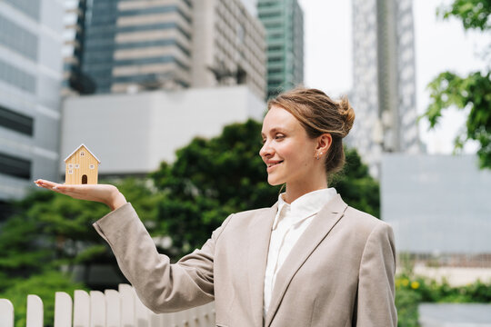 Businesswoman holding small wooden house model on her palm in front of a modern glass office building, real estate investment, property and home ownership