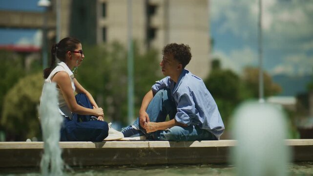 Two school friends are conversing beside a fountain in an urban setting. They are enjoying a casual conversation outdoors on a bright day.