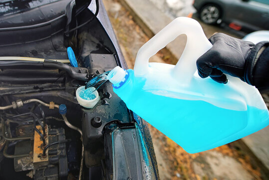 Gloved hand pouring vibrant blue winter screen wash into car washer fluid reservoir to prepare vehicle for freezing weather, safe driving on icy roads. Refilling car windshield washer fluid reservoir