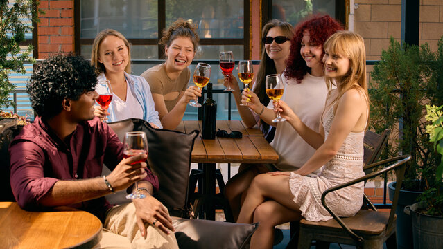 Cheerful company raising wine glasses around sunny terrace table. Concept of collective joy, friendship, shared occasions, summer hospitality and wine campaigns.