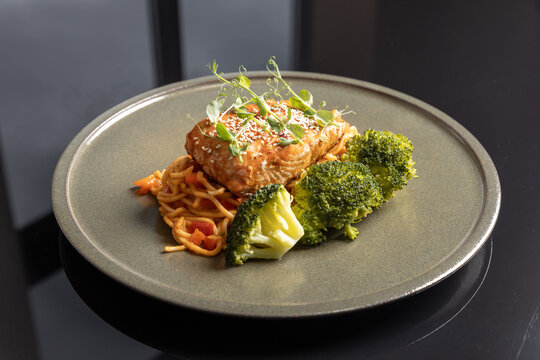 Grilled salmon fillet with noodles, steamed broccoli, sesame seeds, and microgreens served on a modern plate