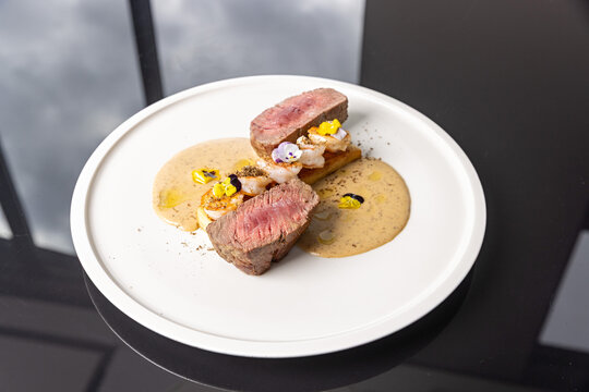 Tenderloin beef steak served with seared prawns, potato terrine, creamy sauce, and microgreens on white plate