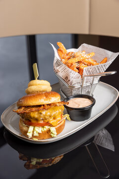Juicy bacon cheeseburger topped with fried onion rings served with French fries and sauce on black reflective table