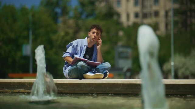 A focused young man is seen studying and reading a book outdoors in a sunny park. He thoughtfully concentrates on his learning while sitting by a peaceful fountain.