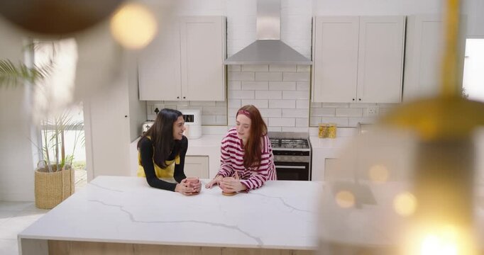 Diverse female friends arriving at kitchen island in yellow dress, sipping pink mugs, catching up