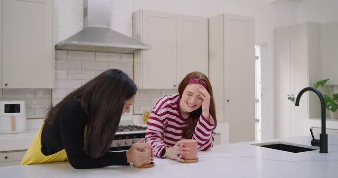 Diverse female friends chatting after Indian woman leaning in at kitchen island over mugs