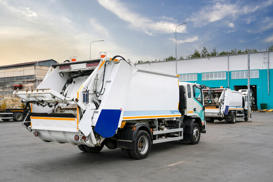 Early morning light casts a warm glow on garbage trucks waiting at a busy waste management facility, ready for the day's collection duties