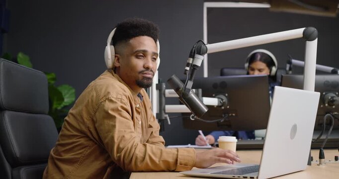 African American man in tan jacket typing, adjusting mic and headset, smiling at studio camera