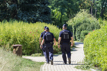 Trzech policjantów patroluje parkową alejkę, spacerując wśród zieleni i zadbanych krzewów. © Henryk Guziak