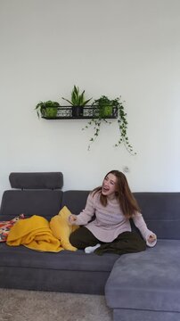 Vertical screen joyful woman jumps with excitement then drops onto sofa laughing and celebrating a happy moment at home expressing energy fun freedom and cheerful emotion