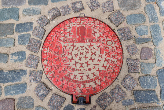 Riga, Latvia - June 24, 2026: Red Rigas Udens utility cover in cobblestone pavement in Riga