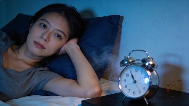 Young Woman Relaxing Awake in Bed at Night with Blue Bedding and Classic Alarm Clock Representing Restful Sleep and Nighttime Routine