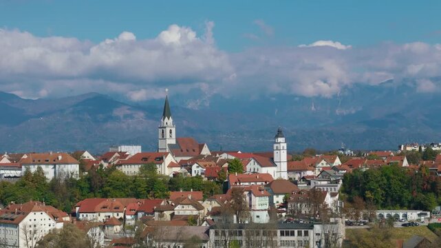 Aerial view shows Kranj, Slovenia with its medieval old town, church towers, and surrounding landscape. Clear blue sky highlights the scene.