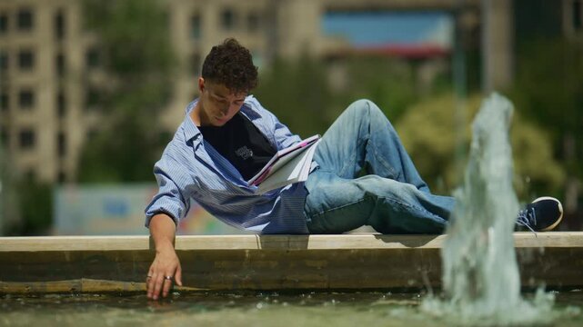 A young man lies comfortably by a city fountain, engrossed in reading notes. He enjoys a peaceful and relaxing moment of study or leisure.