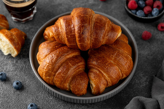 Fresh golden homemade croissants for breakfast with fresh berries and coffee on a gray plate on a dark concrete background.