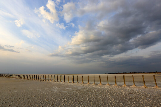 Wooden posts line a vast salt lake under a dramatic cloudy sky at sunset