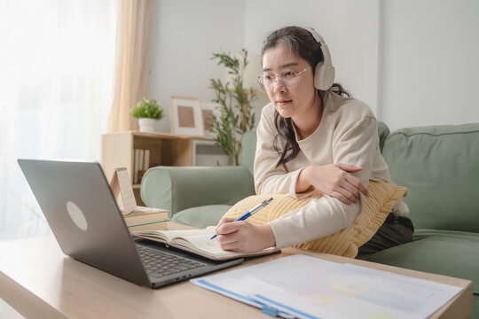 Asian woman wearing headphones writing in notebook while studying online course on laptop at home, concept of e-learning and distance education