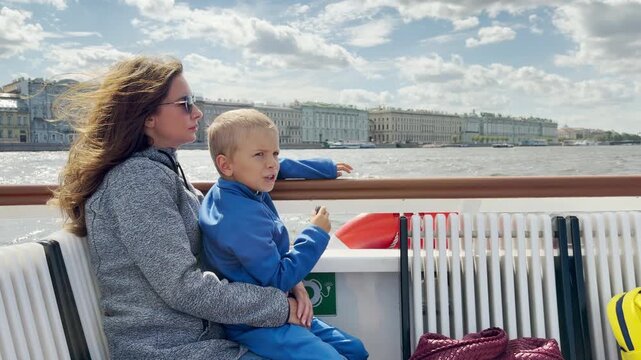 A woman holds a young boy as they sit on a boat along the Neva River in Saint Petersburg. The Winter Palace stands across the water under a cloudy sky. This ride brings a warm, joyful feeling.