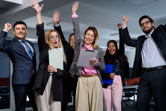 Group of people celebrating in an office during a work event in the afternoon with raised hands and smiles while holding documents and clipboards