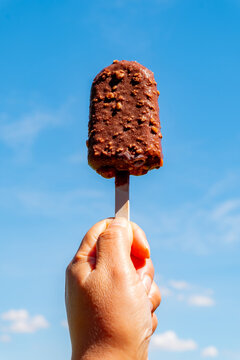 Hand holding crisp chocolate ice cream bar with nuts on wooden stick against clear blue sky in summer. Delicious cold sweet dessert snack outdoors on bright sunny day. Vertical
