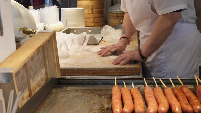 worker rolling dough for dumplings at food market in china. cook preparing dough pieces on board beside sausages. concept of dough craft and street cooking tradition in asia