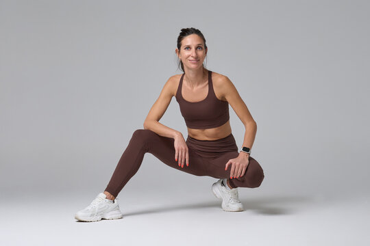 Smiling fit woman in brown sportswear crouching on floor against gray background