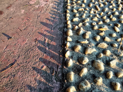 A reflexology stone path embedded in concrete alongside a worn pavement edge, bathed in low angle sunlight that casts long, repeating geometric shadows