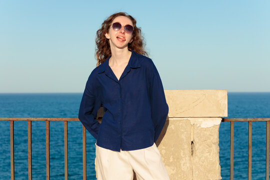 Playful woman with curly hair make tongue out and sunglasses enjoys a sunny day by the sea