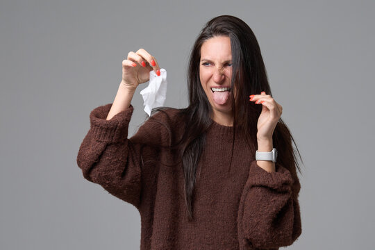 Woman looking at dirty used tissue with disgust showing tongue isolated on grey background